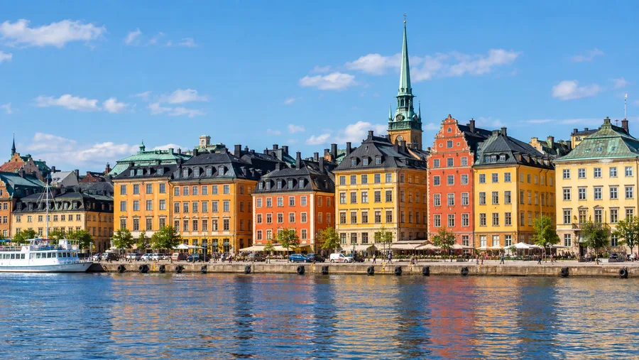 De ockragula, orangea och röda köpmanshusen i Gamla stan sedda från vattnet längs Skeppsbron, en kyrktorn bakom.