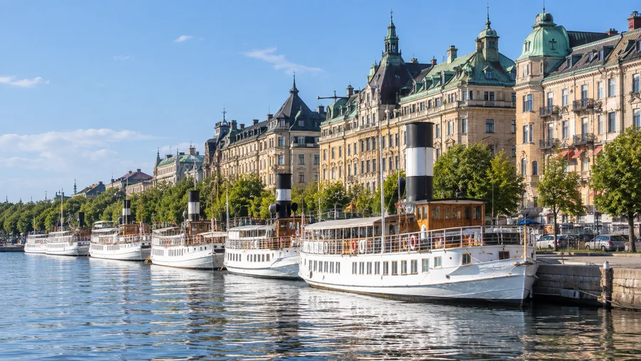 Klassiska vita skärgårdsångbåtar förtöjda längs en stenkaj i centrala Stockholm en sommarmorgon.
