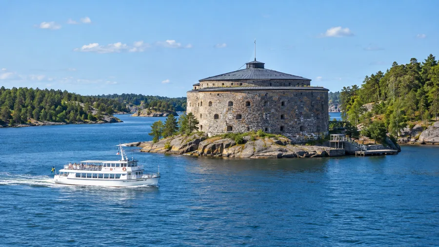 En rund stenfortifikation på en liten klippig ö i ett trångt sund, en vit passagerarbåt som passerar